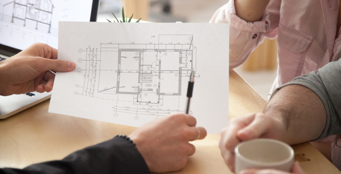 A couple, sat down at a work desk. Being shown printed building structure plans with an architect.