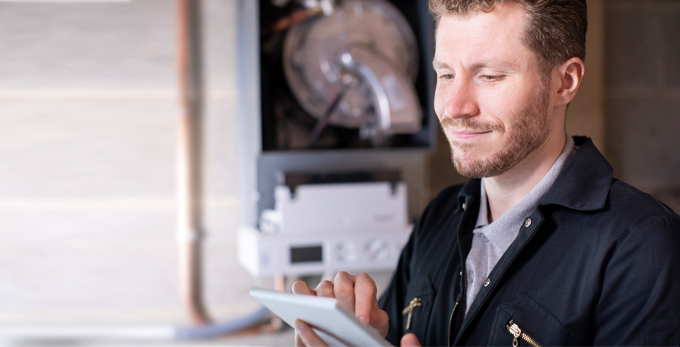 A tradesman holding and looking at a digital tablet. With a boiler in the background with the cover off, showing wiring and vent.
