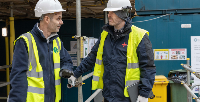An NICEIC approved contractor, talking with another contractor on site. Wearing a safety hi-viz jacket and hard hat, standing on scaffolding.