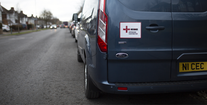 A grey ford transit vehicle, with branded NICEIC labels and license plate. Parked up stationary on a street, outside of a client household.