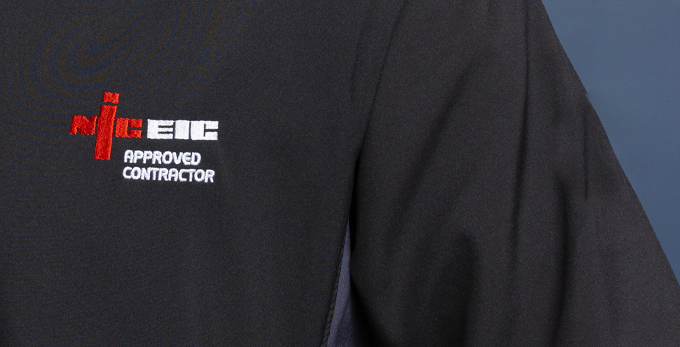 A close up of a black NICEIC branded polo shirt in a kitchen with a blue cladded background.