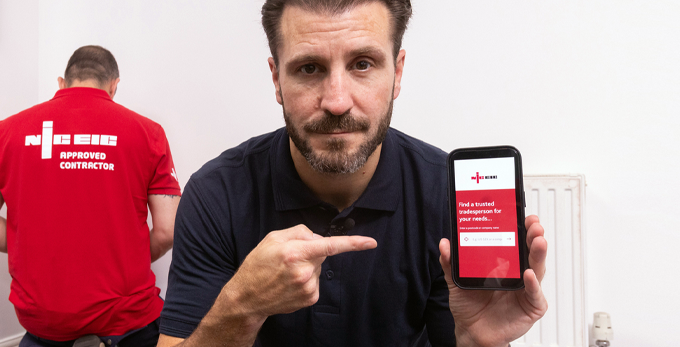 Male in household holding mobile phone, showcasing website to find a trusted tradesperson. Whilst tradesman in red polo shirt undertakes electric work.