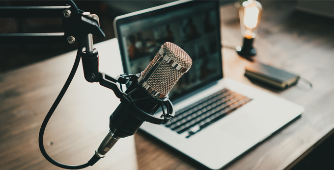 A close up of a podcast microphone, with a apple Mac book computer on the deck in a podcast studio.