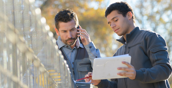 two contractors looking at a digital tablet, whilst one person is on a mobile phone, outside on a construction site.