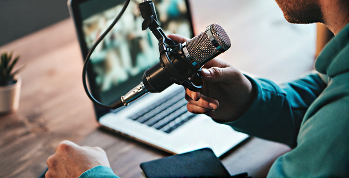 A male smiling, in a green hoodie, conducting a podcast whilst sat at a clean deck, using a apple Macbook pro laptop.