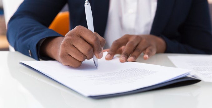 A person, sat at a deck reading a paper contract, holding a pen to sign and date.