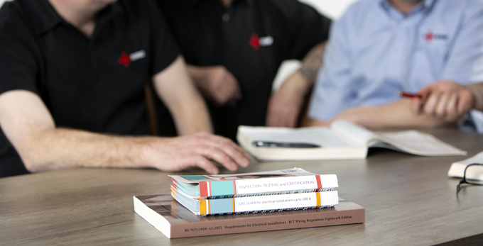 Three contractors are communicating and making notes whilst attending an NICEIC training course. There are training and learning documents stacked up on the contractors table for reference. 