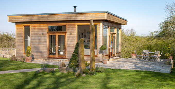 A wide view shot of a modern garden office on a bright summers day, a clear sky is above the wooden office and standing stones can be seen on the grass.
