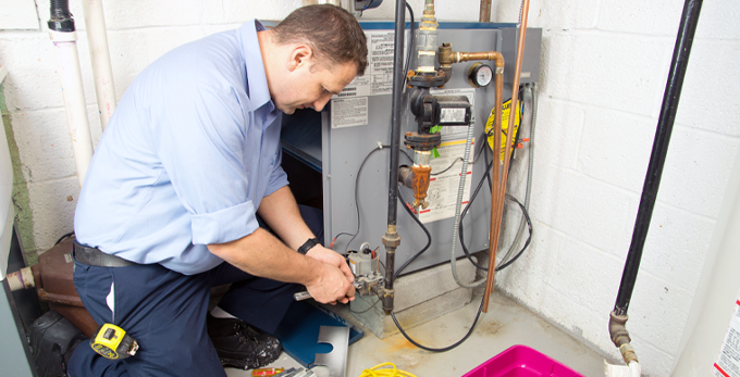 A plumber wearing a smart light blue shirt, kneeling down, fixing a gas furnace.
