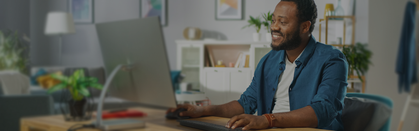 A young male wearing a blue shirt, happily working at home in a modern living room, sat at a computer desk.