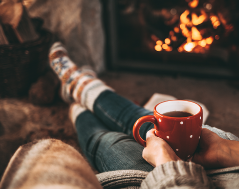 Woman in woollen socks by the fireplace. Unrecognisable relaxes by warm fire with a cup of hot drink and warming up her feet in woollen socks. Cozy atmosphere. Winter and Christmas holidays concept.