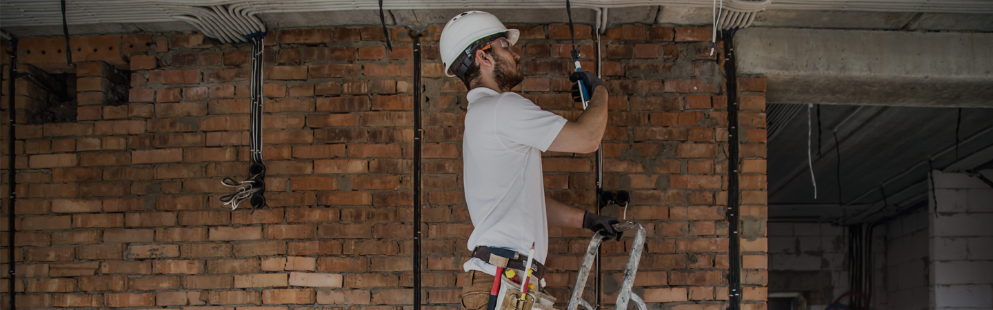 An electrician standing on a step ladder, working on second electrical fix components, including plug sockets and light fittings on a building site.