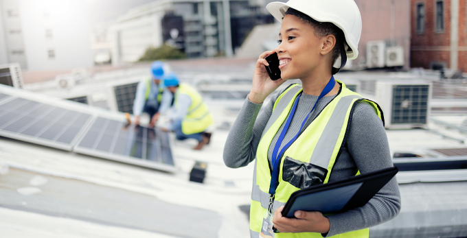 A team of two installing solar energy panels on a roof, whilst the site manager is happily communicating on a phone.