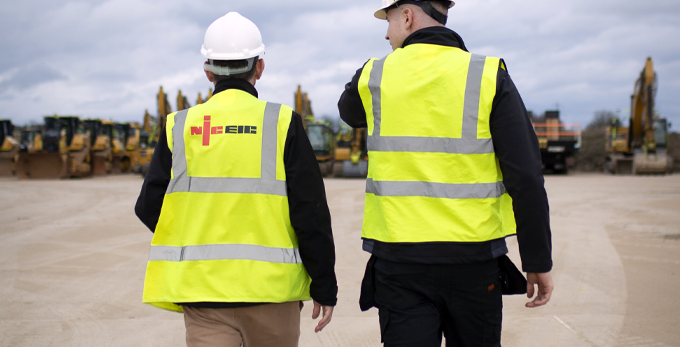 Two NICEIC consultants, wearing protective safety gear and branded high-viz jackets, walking towards a construction site.