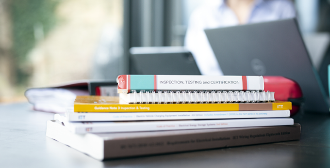 Close up of multiple learning and qualification documentations stacked up on an office table. Including inspecting, testing and certification for electricians.