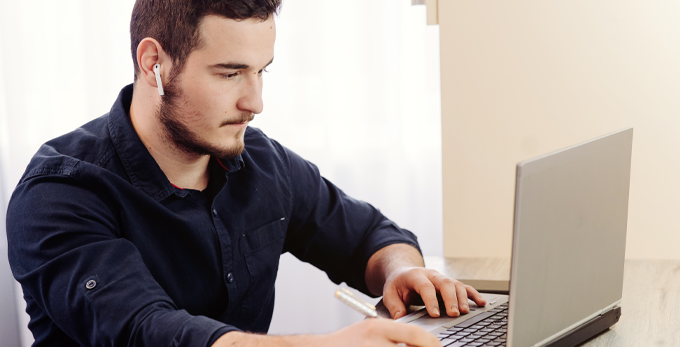 Male working on computer remotely, whilst making notes in a book sat down at a deck.