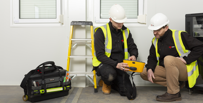 Two NICEIC approved contractors on site at a customers household, wearing a hard hats and hi-viz jackets, kneeing down reviewing data on an electrical reading device.
