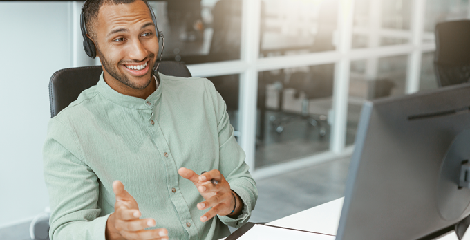 Young male wearing light green shirt, sat down at deck communicating on a computer, in an office building.