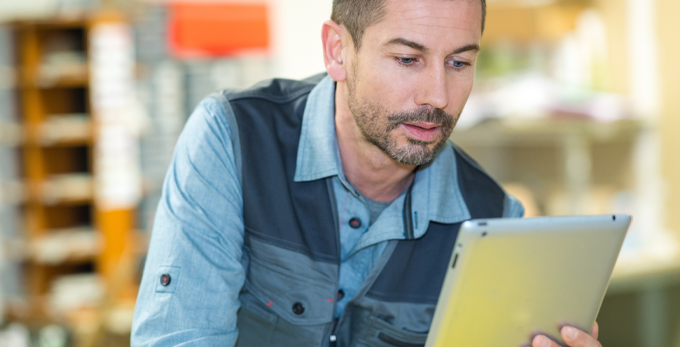 A tradesman wearing protective clothing, holding and looking at a grey digital tablet whilst in an office.