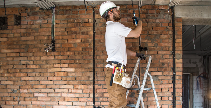 An electrician standing on a step ladder, working on second electrical fix components, including plug sockets and light fittings on a building site.