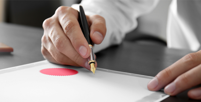 A male seated at an office deck, holding a black fountain pen, ready to sign a printed certificate.