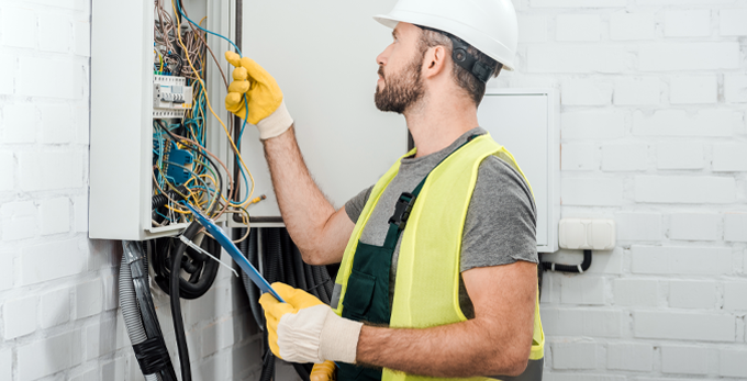 Electrician holding clipboard and checking the wiring in electrical board in corridor.